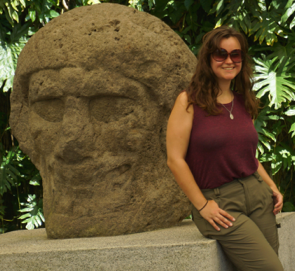 Image of a White woman wearing a tank top and sunglasses next to a stone statue of a head. 