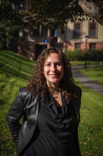 White person who presents as feminine, with curly brown hair, stands in front of a building and lawn. They're wearing a black leather jacket and black blouse. 